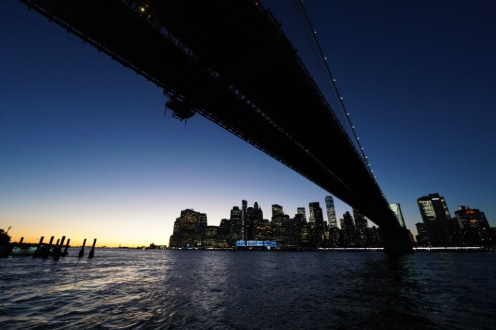 Wide angle photo taken below the Brooklyn bridge, on the Brookly side. The underside of the bridge is visible across the photo, and the financial district is visible in the distance across the water. The photo is taken at dusk.